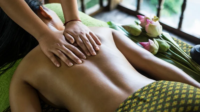 Woman lying face-down on a massage table covered with a green cloth receiving a back massage from a masseuse using both hands